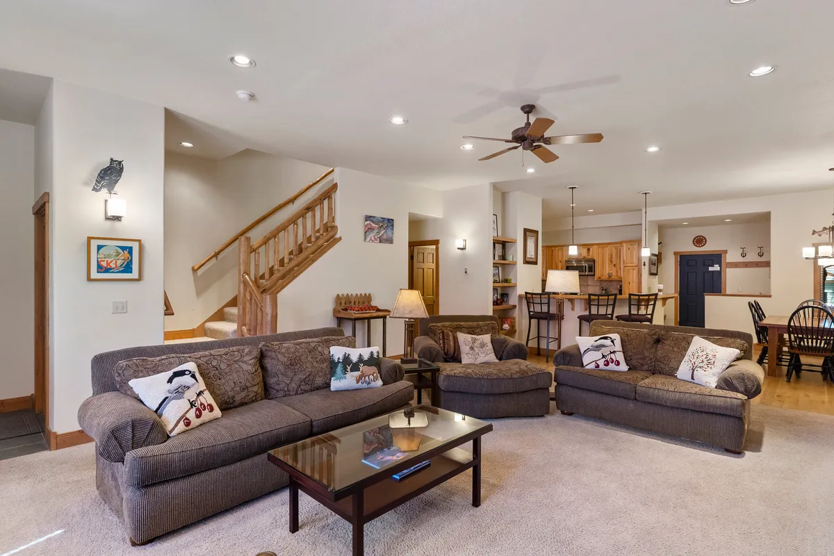 The image shows a warmly lit, inviting open-concept living space with plush sofas arranged around a coffee table, a stylish staircase, and a spacious dining area nearby. The ambiance is cozy, perfect for relaxing or gathering with loved ones after a day on the slopes. The space hints at evenings spent by the fireplace and shared meals around the table, ideal for families or small groups.