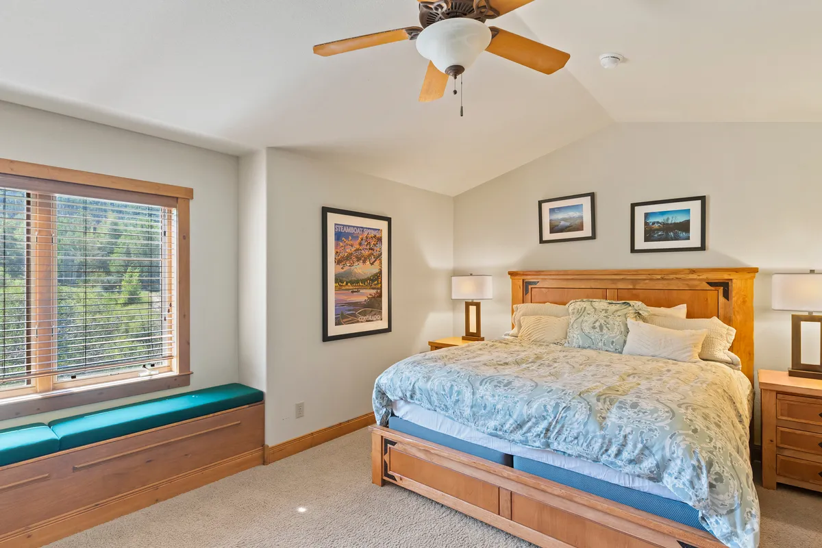 A serene main bedroom featuring a king-size bed with plush bedding, large windows that let in natural light, framed artwork above the bed, and nightstands with lamps. The inviting ambiance emphasizes comfort and relaxation, with mountain views visible through the windows.