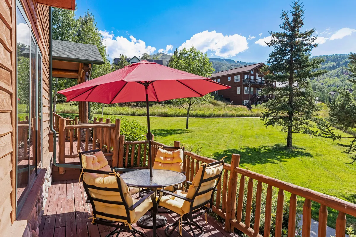 A tranquil green lawn with a table and chairs under a red umbrella sits before a stunning mountain backdrop. The scene evokes peaceful mornings, outdoor dining, and a connection to nature. The space hints at neighborhood walking paths and opportunities for wildlife viewing.