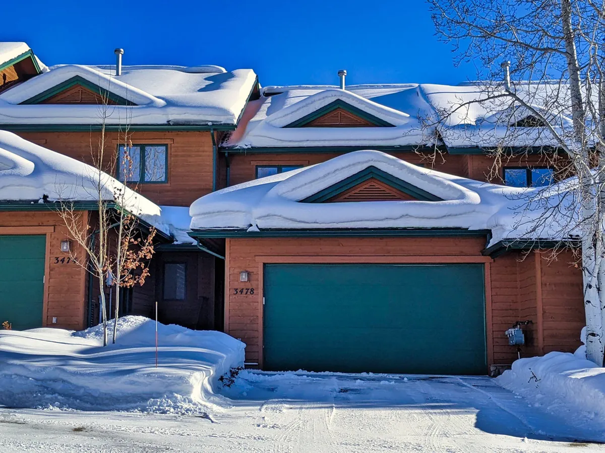 This image captures the home's exterior during winter, prominently featuring the green garage doors, wooden siding, and aspen trees in a tranquil, snowy setting. The garage is clearly visible, suggesting convenient storage for outdoor gear and easy access after mountain adventures.