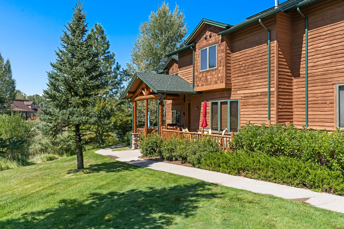 A welcoming exterior view of the property featuring a wooden facade and a porch with vibrant red accents. Lush greenery surrounds the home, and a pathway leads invitingly toward the house. The porch offers space for relaxing and enjoying the natural setting.