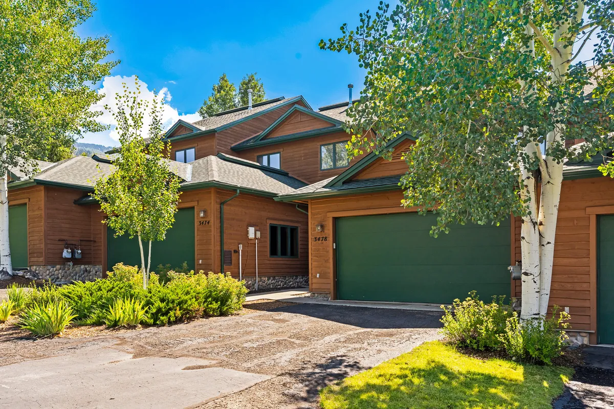 The front exterior of the home showcases the green garage doors, landscaped garden, wooden siding, and aspen trees. The driveway is visible, highlighting extra parking space and the home's welcoming atmosphere in a lush, green season.