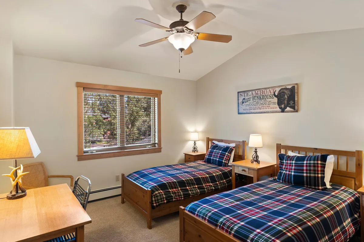 A bright and functional twin bedroom with two comfortable twin beds, wooden furniture, a desk with chair, lamp, and a window providing natural light. The desk area is ideal for kids’ activities or remote work, supporting flexibility for guests.