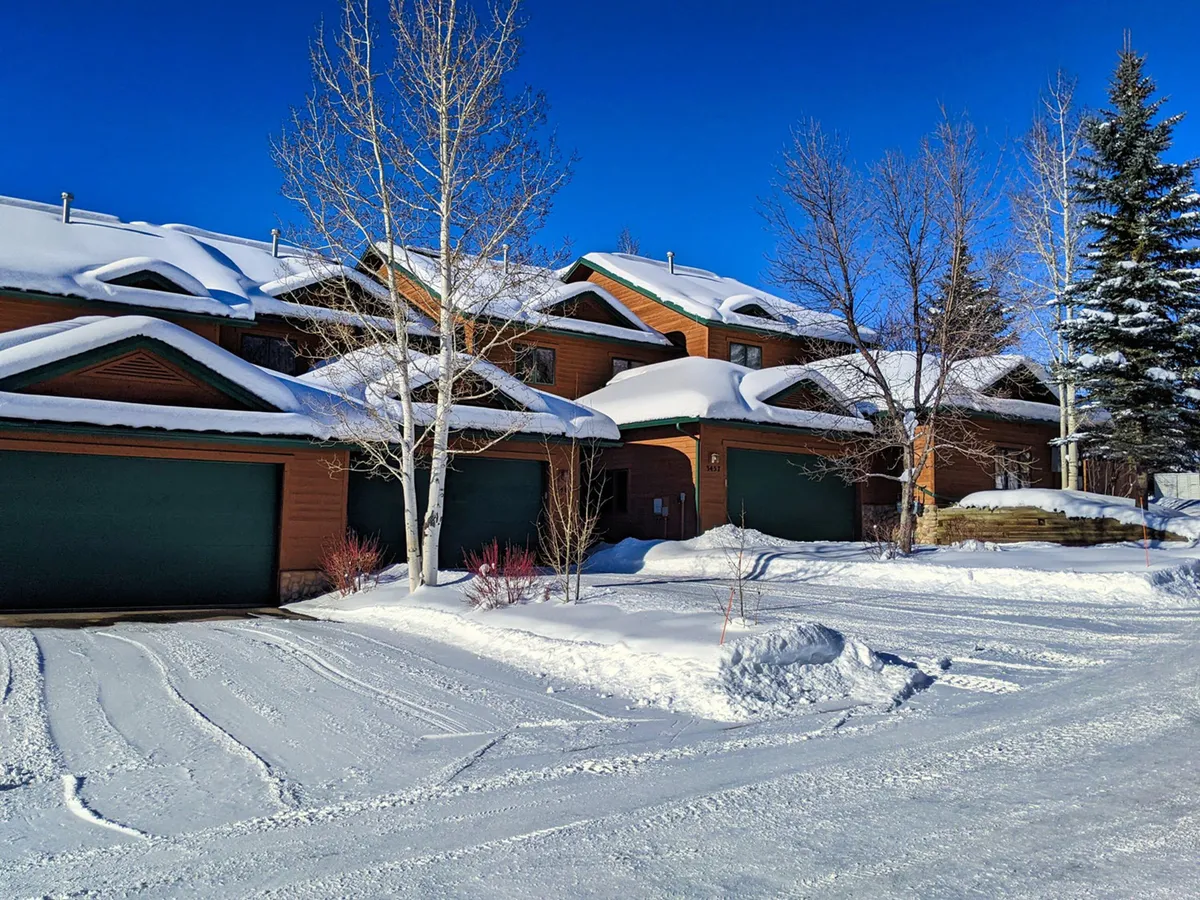 A serene winter exterior view of the property, with snow-covered roofs, wooden exteriors, and green garage doors. The landscape is blanketed in white snow, evoking a classic ski destination atmosphere. The image suggests easy access to outdoor winter activities and après-ski relaxation.