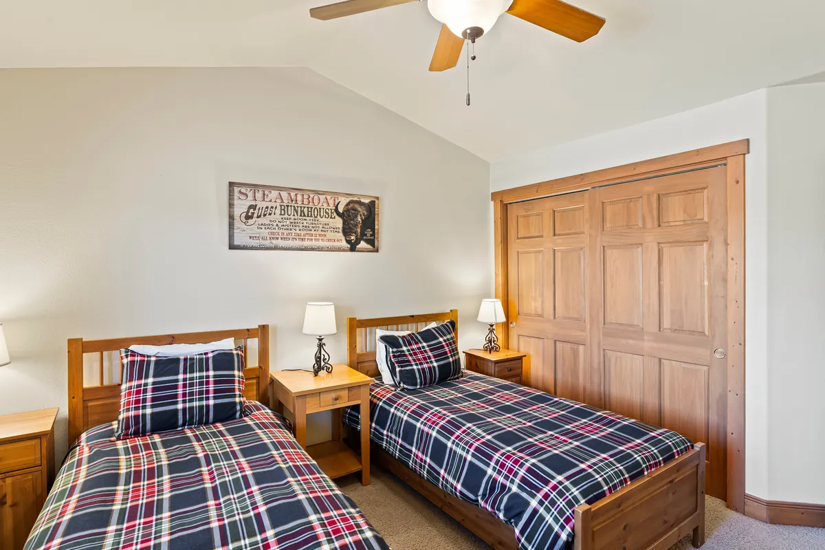 A cozy interior scene with plaid bedding, wooden furniture, and art on the walls. Sunlight streams through the window, casting a warm glow and emphasizing a relaxing spot to start or end an adventurous day.