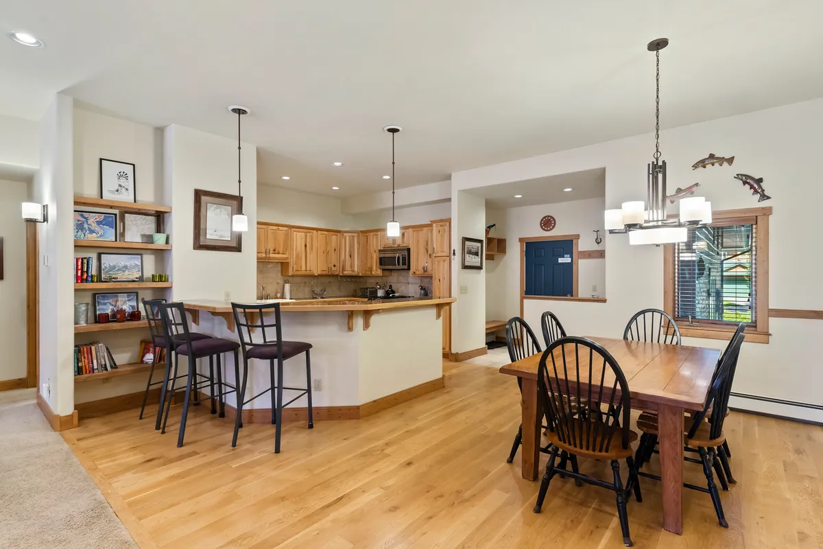 A bright and spacious dining area featuring a large dining table, a kitchen island, and a bookshelf, all set within an inviting open-concept layout. The scene suggests space for family meals, socializing, and shared activities.